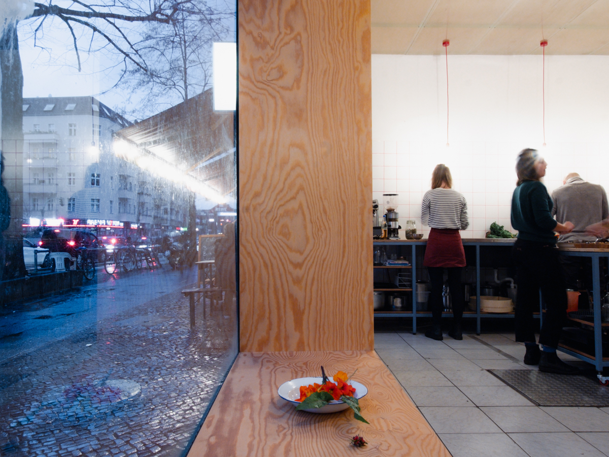 Blick in Fenster: Linke Seite Gehweg und Straße, rechte Seite Innenraum mit Blick auf Küche. Personen stehen und hantieren in Küche. Im Vordergrund: Teller mit Blüten.