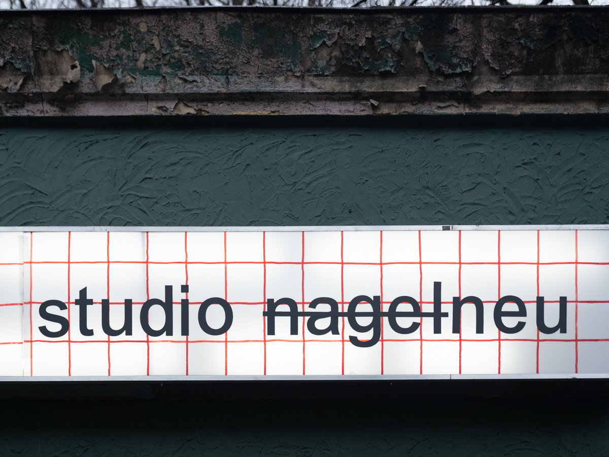 Helles Schild mit Karo-Muster von dunkler Wand. Schriftzug sagt studio nagelneu.