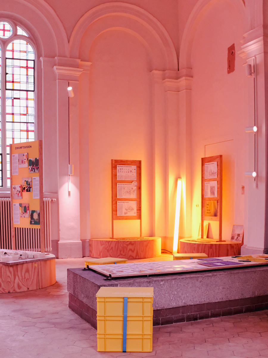 Rosa beleuchteter Ausstellungsraum in Kirche mit Podesten und Hocker.